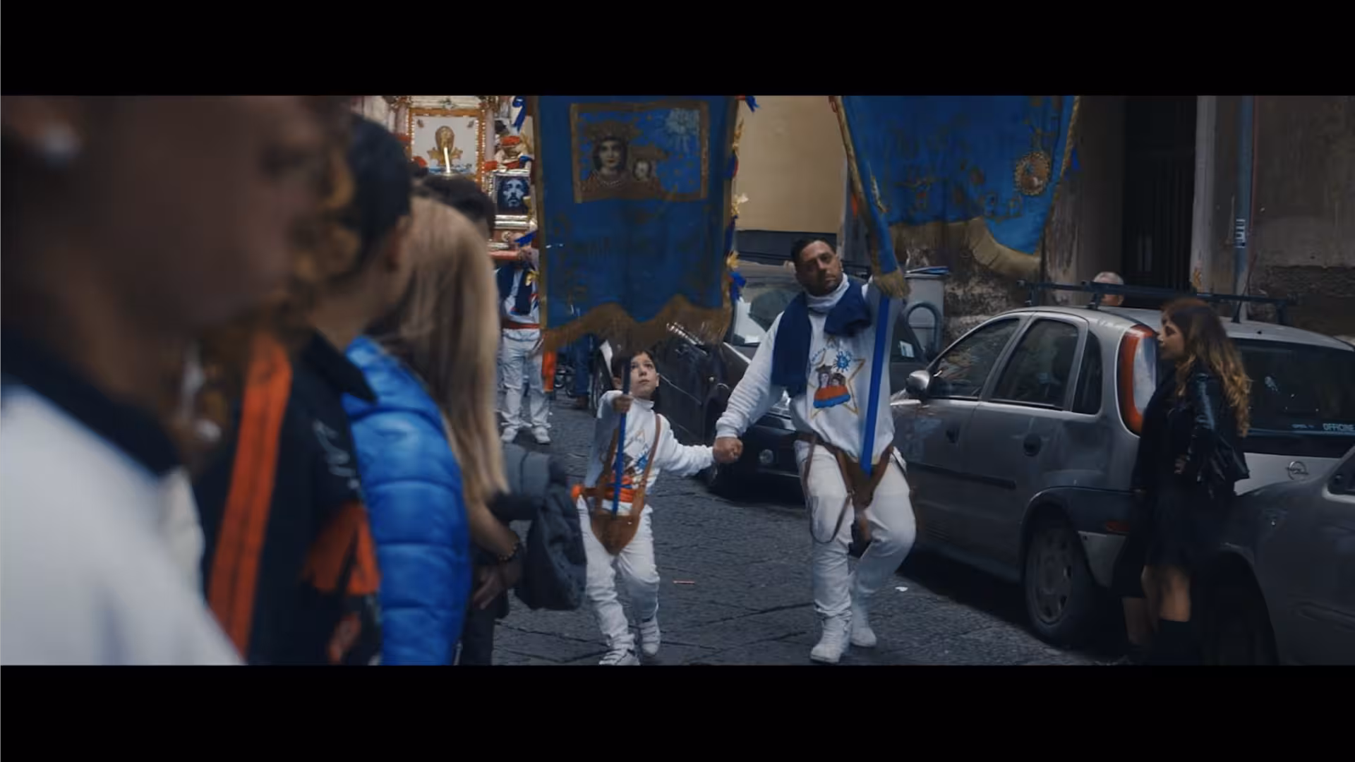 A religious procession; a father and son, dressed all in white, carry blue banners with religious icons on them.